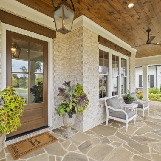 Front porch of a home with stone flooring, featuring a wooden door and lantern above. Two gray chairs sit by large windows, flanked by potted plants. A ceiling fan is visible above, and another house is seen in the background.