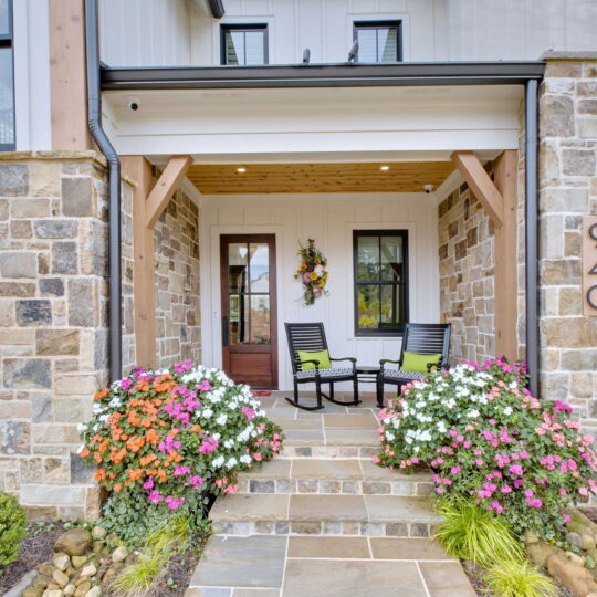 Front porch of a house with two black chairs and a small table. Colorful flowers in planters line the stone steps. The house features stone siding and wood accents. The number 940 is mounted on the wall.