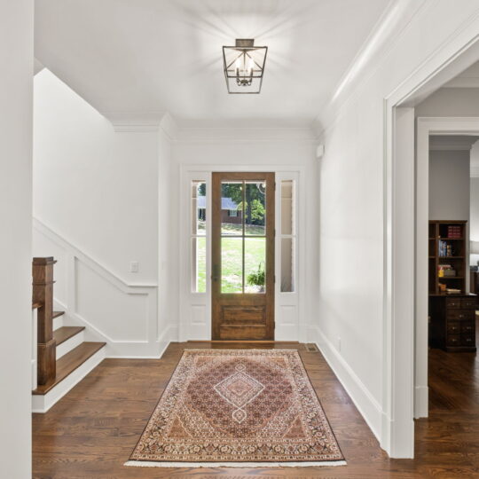 A bright entryway with wooden flooring and a decorative rug. There is a wooden glass-paneled front door, stairs on the left, and a doorway on the right. A geometric ceiling light fixture hangs above.