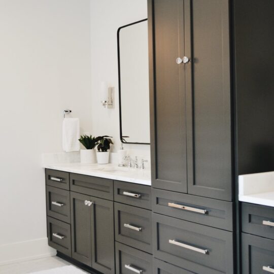 Modern bathroom with dark gray cabinets featuring multiple drawers. A white countertop supports a sink with a chrome faucet. Above is a rectangular mirror. The floor is light-colored tile with a white bath mat. A potted plant and towel are on the counter.