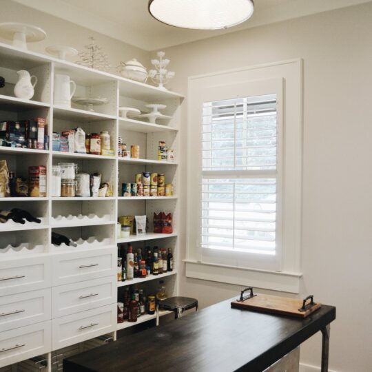 A well-organized pantry features open shelves stocked with various food items and a dark wooden table with a cutting board. A large black pendant light hangs above, and a window with blinds lets in natural light, illuminating the brick floor.