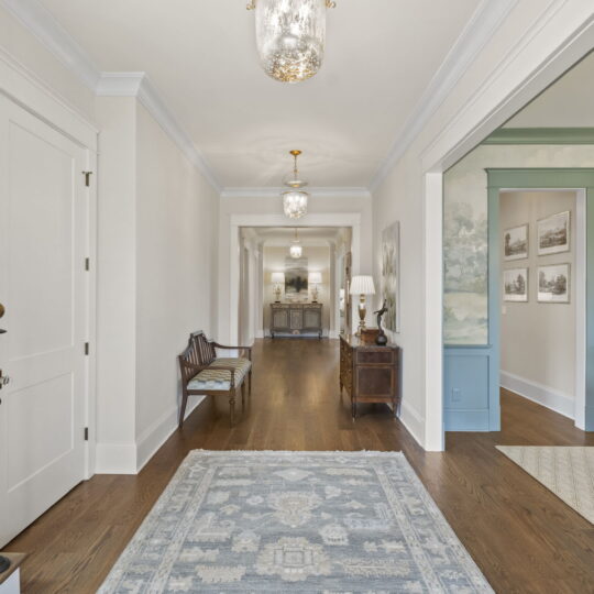 A long, elegant hallway with polished wooden floors, adorned with a patterned blue rug. It is lit by ornate ceiling lights. The walls are decorated with framed artwork, leading to a distant mirrored sideboard and a warmly lit interior.