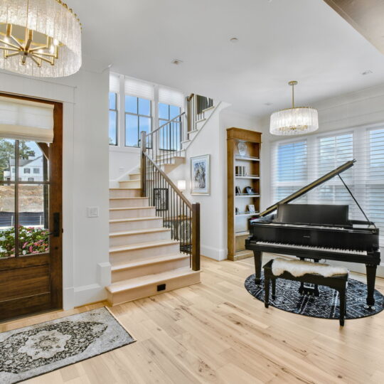 A bright living room with a grand piano on a circular rug. To the left, a wooden door with glass panels and floral decor. A staircase with wooden steps leads upward. The room has large windows with blinds and two chandeliers for lighting.