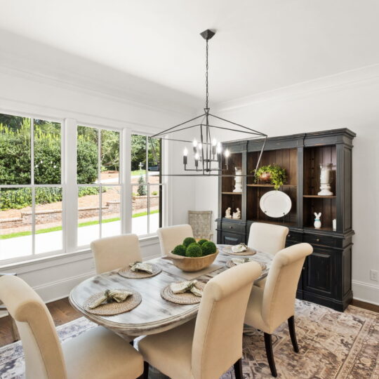 A dining room with beige upholstered chairs around a wooden table set with plates and decor. A black china cabinet displays decorative items. A geometric chandelier hangs above, and large windows bring in natural light. The floor is dark wood with a rug.