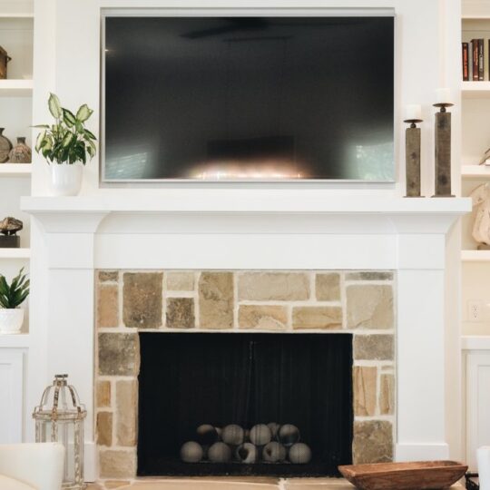 A cozy living room features a stone fireplace with a television mounted above it. The mantel is decorated with candles, and built-in shelves on either side hold books, plants, and decorative items. Neutral tones create a serene atmosphere.