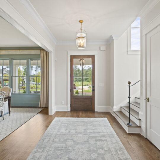 A bright and elegant home interior featuring a wooden front door with glass panels, a formal dining room with a chandelier on the left, and a staircase on the right. Large windows offer a view of the greenery outside.