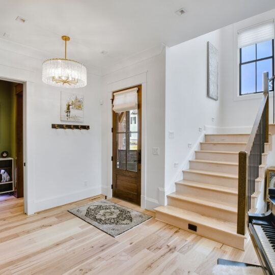 A bright hallway featuring light hardwood floors and white walls. A staircase with white risers and wooden steps leads up. A chandelier hangs from the ceiling. On the right is a black piano, and an open doorway on the left reveals a green room.