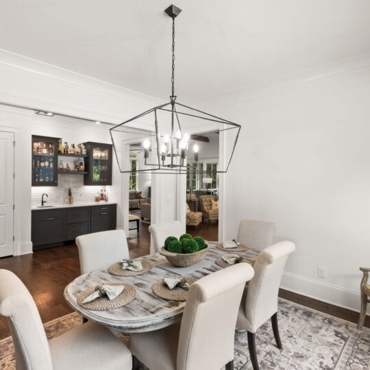 A dining room with a round marble table set for six, surrounded by beige upholstered chairs. A geometric chandelier hangs overhead. A cabinet and counter with a sink are in the background. Wood flooring and a patterned rug are visible.