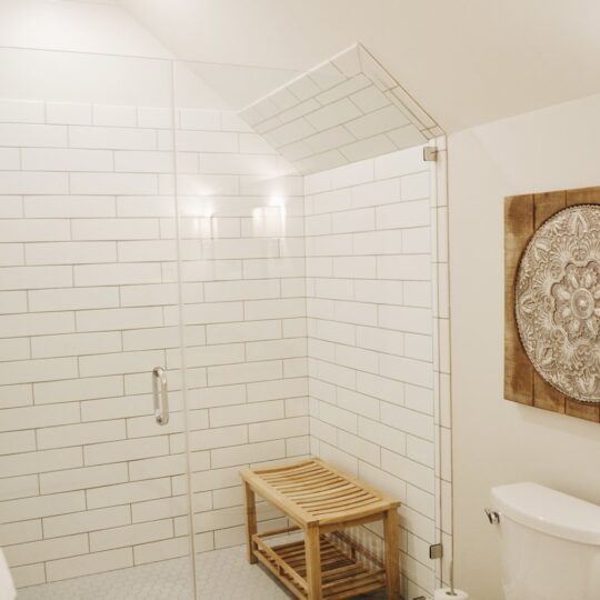 A clean bathroom featuring a glass-enclosed shower with white subway tiles and a wooden bench. The floor tiles are patterned, and theres a white toilet on the right. A carved decorative panel hangs on the wall.