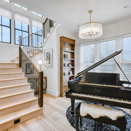 A bright room featuring a grand piano on a round black rug, a staircase with wooden steps and black railings, a large window with blinds, and a chandelier. A bookshelf and artwork adorn the white walls.