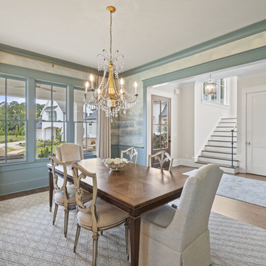 A dining room with a wooden table and six chairs, a chandelier above, large windows with light curtains, and a view of a staircase leading to another level. The walls have a subtle landscape mural, and there is a blue area rug by the stairs.