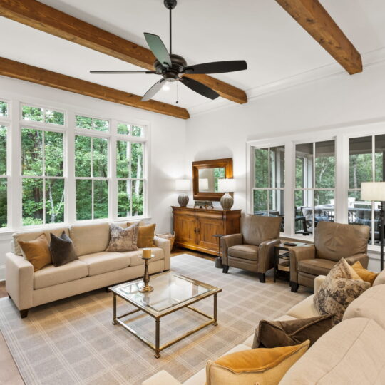 A cozy living room with beige sofas, a glass coffee table, and armchairs facing each other on a patterned rug. Large windows reveal a lush green view. Exposed wooden beams on the ceiling and a ceiling fan add a warm touch. Lamps and cabinets complete the decor.