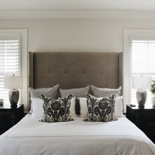 A neatly arranged bedroom with a large bed featuring a gray upholstered headboard and decorative pillows. Two matching nightstands hold lamps and plants. Light filters through the window shutters, creating a serene atmosphere.