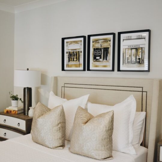 Bedroom with a beige upholstered headboard, three decorative pillows, and white bed linens. Two bedside tables with matching lamps flank the bed. Three framed pictures hang above the headboard, and theres a decorative wall piece on the left.