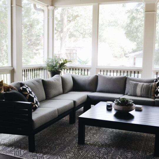 A cozy screened-in porch with a gray sectional sofa, black and white patterned cushions, and a cat lounging on the corner. A black coffee table with decorative items sits on a patterned rug. Lush greenery is visible through the screens.