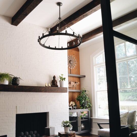 A cozy living room with a white brick fireplace, wooden mantel, potted plants, and Buddha statue. A round chandelier hangs from the ceiling, and large windows on the right let in natural light, illuminating a chair and small table in the corner.