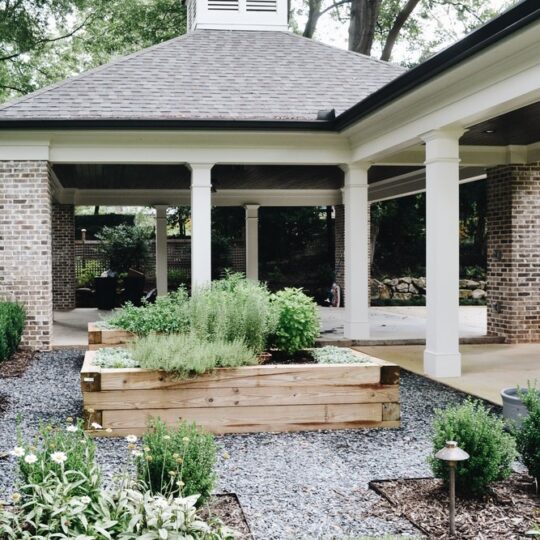 A garden scene with a wooden raised bed full of lush greenery. Its surrounded by gravel and various plants. In the background, theres a covered area with brick pillars and a wooden roof, set amidst a tranquil environment.