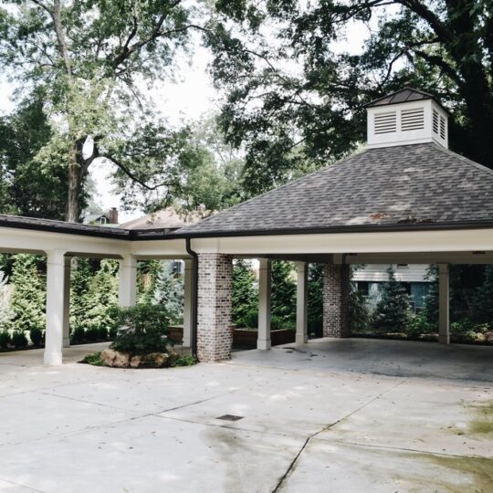 A covered carport with a gable roof supported by white columns and brick pillars in a lush garden setting. The area is shaded by large trees, with a concrete driveway leading to the structure.