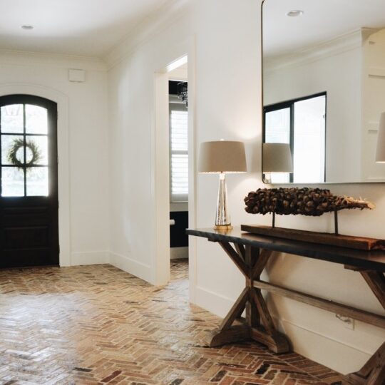 A hallway with herringbone-patterned brick flooring, featuring a dark wooden console table with two lamps and a decorative piece. A large mirror hangs above. A dark wooden door with a window and a wreath provides natural light.