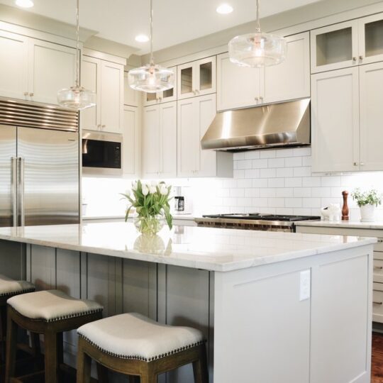 Modern kitchen with light cabinets, a large island with three cushioned stools, stainless steel appliances, and hanging pendant lights. White subway tile backsplash and a vase of tulips on the counter add a touch of elegance.