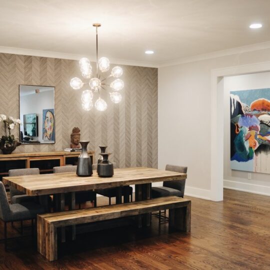 A modern dining room features a wooden table with benches and gray chairs. A stylish chandelier hangs above. The wall displays a large colorful abstract painting and a mirror with a wooden sideboard beneath, adorned with decor and a plant.