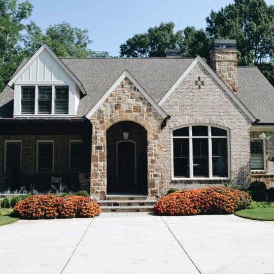 A charming stone and brick house with a gabled roof. The front yard features orange flowers along the walkway and trimmed shrubs. Tall trees surround the property, and the driveway is visible in the foreground.