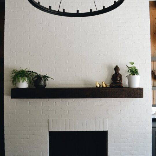 A white brick fireplace with a dark wooden mantle. On the mantle are plants, a Buddha statue, and decorative pieces. Wooden shelves flank the fireplace, displaying various decorations. A circular black chandelier hangs above.