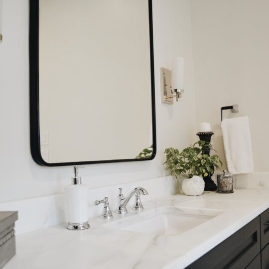 Modern bathroom with a large rectangular mirror, white countertop, and dark cabinetry. A silver faucet is over the sink, with a soap dispenser nearby. A potted plant and a pumpkin decoration add a touch of greenery and seasonal decor.