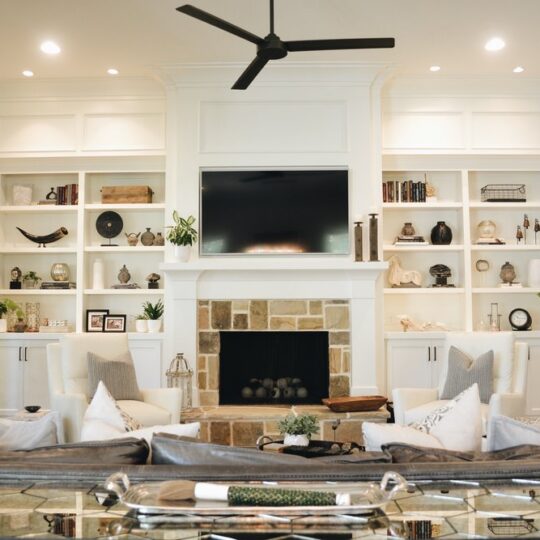 A cozy living room with a white fireplace and built-in shelves filled with decor items. Two plush chairs and a glass table are in front of the fireplace. A ceiling fan hangs overhead, and there are plants on either side of the fireplace.