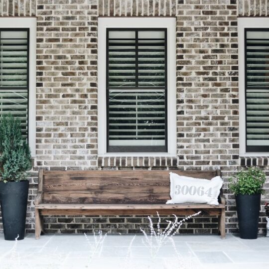 A rustic bench with a decorative pillow sits in front of a brick wall with three windows. Two tall potted plants and two smaller pots are placed on the stone patio, adding greenery to the scene.