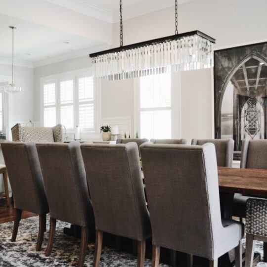 A modern dining room with a long wooden table surrounded by gray upholstered chairs. A rectangular chandelier hangs above. Large windows with white shutters let in natural light. A piece of framed art is on the wall, and bar stools are at a counter.