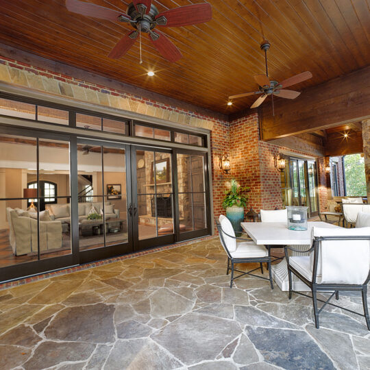 A spacious patio with stone flooring, featuring a dining set with white cushioned chairs. Large sliding glass doors reveal a cozy indoor living area. Ceiling fans are installed on the wooden ceiling, and plants and lights enhance the ambiance.