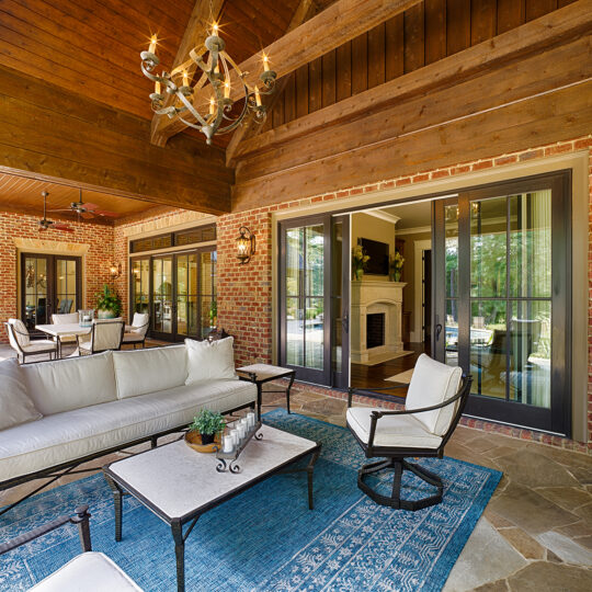 A spacious outdoor patio with a brick wall, featuring a white couch and chairs around a coffee table on a blue rug. Glass doors lead inside to a room with a fireplace. A wooden ceiling with a chandelier and a ceiling fan is overhead.