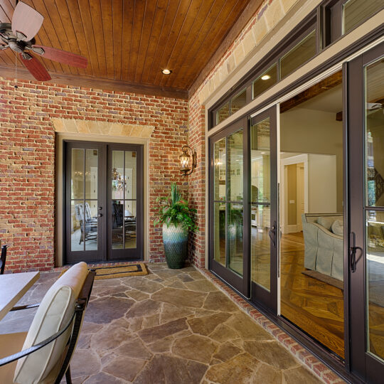 A cozy patio with brick walls and a wooden ceiling features a table with chairs, large glass doors, a ceiling fan, and a potted plant. Natural light streams in, showcasing a glimpse of the greenery outside.