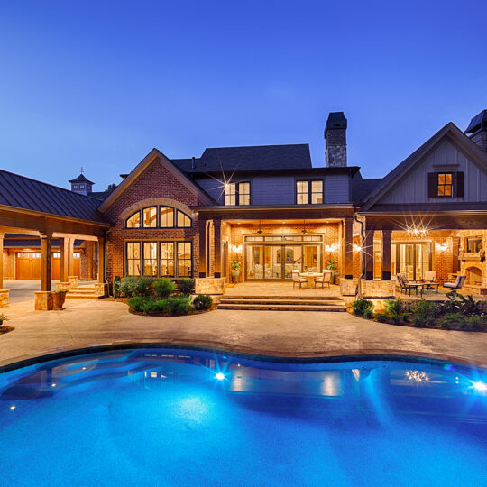 A luxurious mansion is illuminated at dusk, showcasing a grand facade with stone and wood accents. In the foreground, a glowing blue swimming pool reflects the warm lights of the house, surrounded by a spacious patio and greenery.