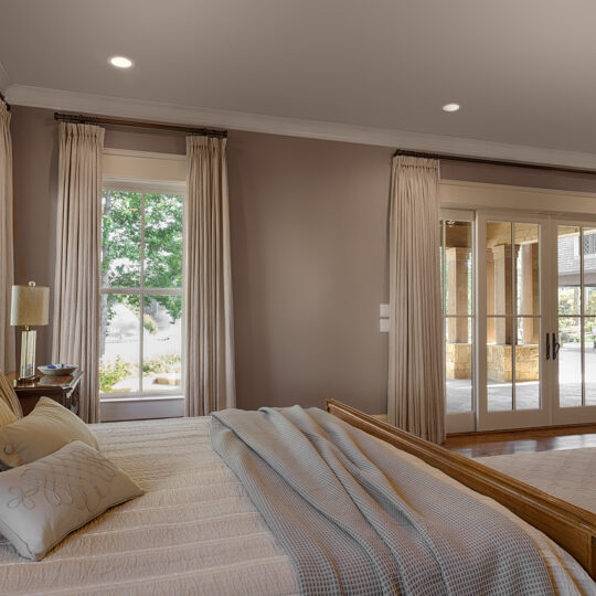 A spacious bedroom with a neatly made bed, light-colored bedding, and decorative pillows. Large windows with beige curtains allow natural light to flood the room. Wooden flooring and glass doors open to a patio with a garden view.