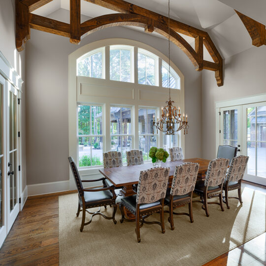 A spacious dining room with a wooden table and eight upholstered chairs on a beige rug. Large arched windows let in natural light. The ceiling features exposed wooden beams, and a chandelier hangs above the table. A clock is on the wall.