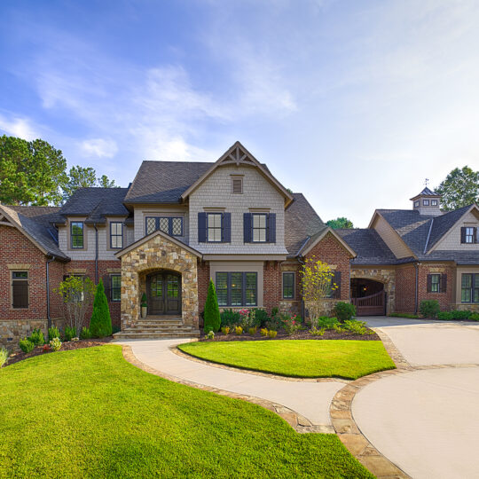 A large, elegant house with a mix of brick and stone facade under a clear blue sky. It features multiple gables, a chimney, and a manicured lawn. The driveway is smooth and curves towards the entrance. Trees are visible in the background.