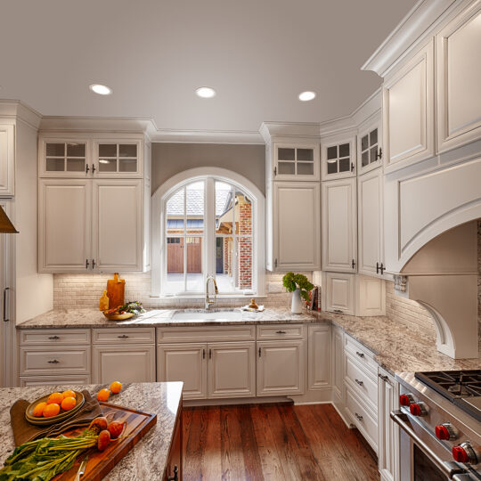 A spacious kitchen with white cabinets, brown granite countertops, and a large arched window. There are fruits on a cutting board, a pot on the stove, and decorative lighting. The floor is wooden, complementing the elegant ambiance.