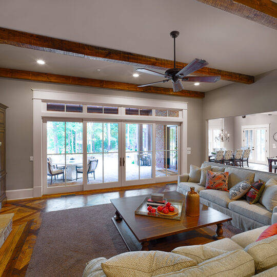 Spacious living room with a beige sectional sofa, a wooden coffee table topped with decor, and a stone fireplace. Large glass doors open to a patio, and wooden ceiling beams add warmth. The adjacent kitchen is visible through an open doorway.