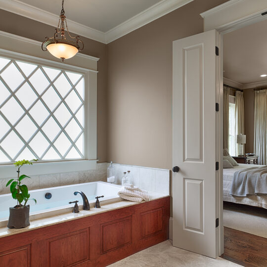 Bathroom with a wooden-framed bathtub featuring a potted plant, adjacent to a bedroom. A large window with a grid pattern allows natural light in, and an elegant chandelier hangs from the ceiling. The bedroom has a bed and is softly lit.