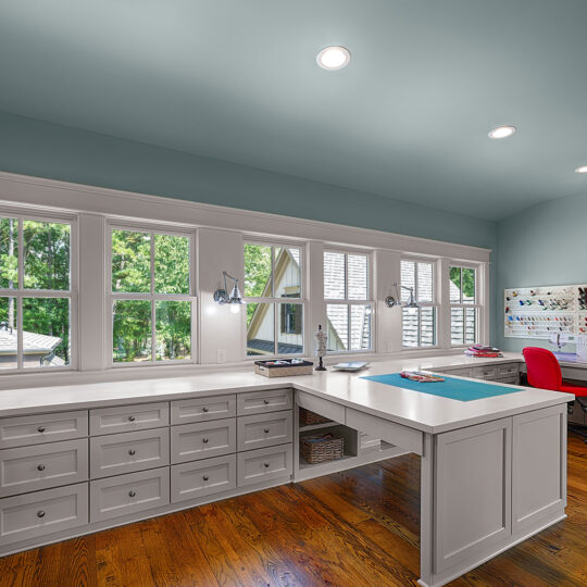 Spacious sewing room with large windows and hardwood floors. White desks with drawers are arranged in a U-shape. A red chair sits at a desk, and spools of thread are organized on the wall. Trees are visible outside the windows.