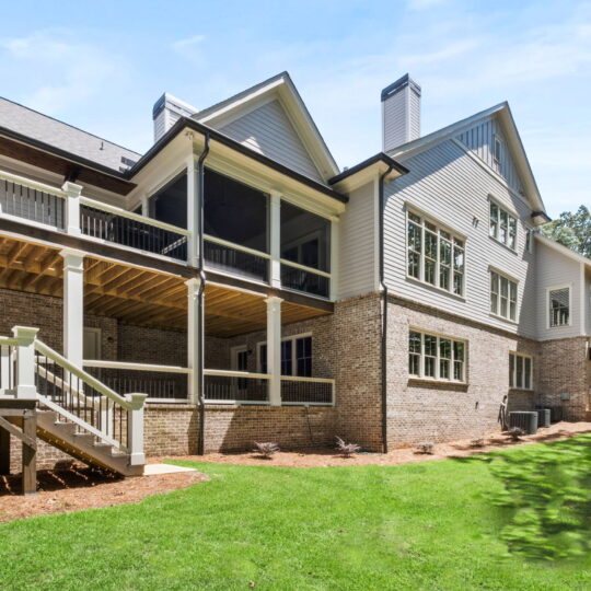 Backyard view of a large modern house with multiple stories. The facade features a mix of brick and siding, with several windows and a covered balcony. A staircase leads down to the green lawn, surrounded by trees and landscaping.