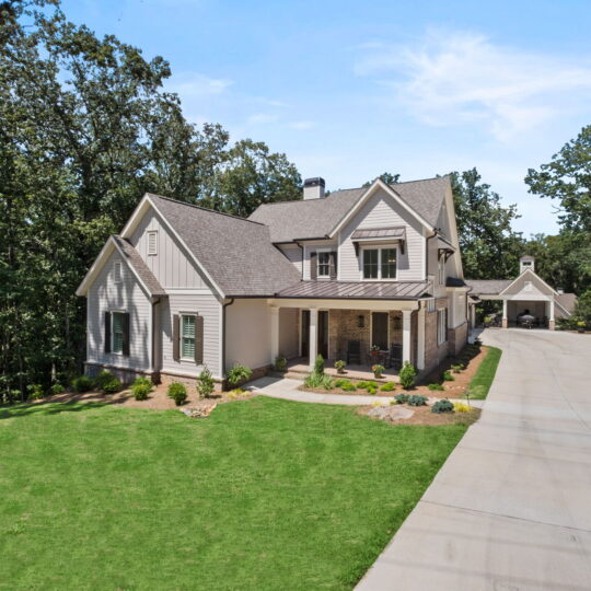 A large two-story house with a gabled roof and a porch, surrounded by trees. It features a spacious driveway and well-manicured lawn. The sky is clear and sunny.