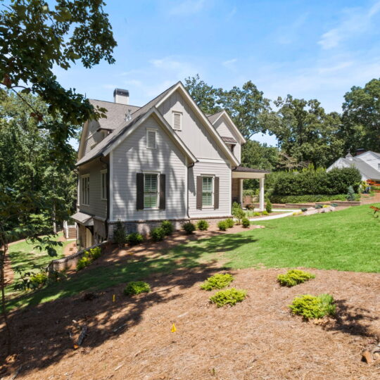 A light gray, two-story house with a gabled roof is surrounded by a well-maintained lawn and trees. A smaller house is visible in the background, nestled among greenery under a clear blue sky.