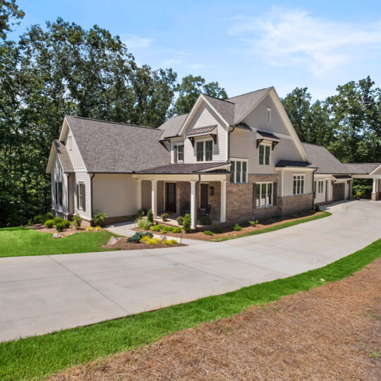 A large, modern two-story house with gray siding and a gabled roof. It features a spacious driveway, landscaped front yard, and is surrounded by trees. The garage is detached and connected by a covered walkway.