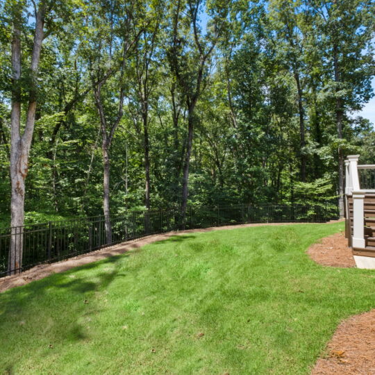 A lush, green backyard with a wooden staircase leading down from a porch. The yard is bordered by a black metal fence and dense trees, creating a serene and natural setting under a clear blue sky.