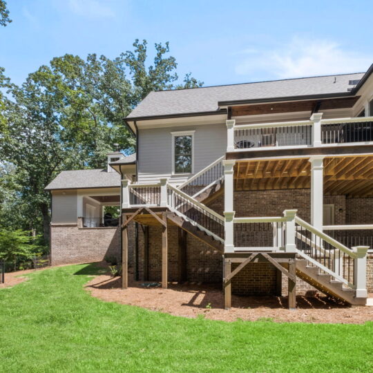 A multi-level house with beige siding and brick foundation, featuring a wooden deck and staircase. The yard has lush green grass and is bordered by tall trees. A black fence runs along the left side under a clear blue sky.