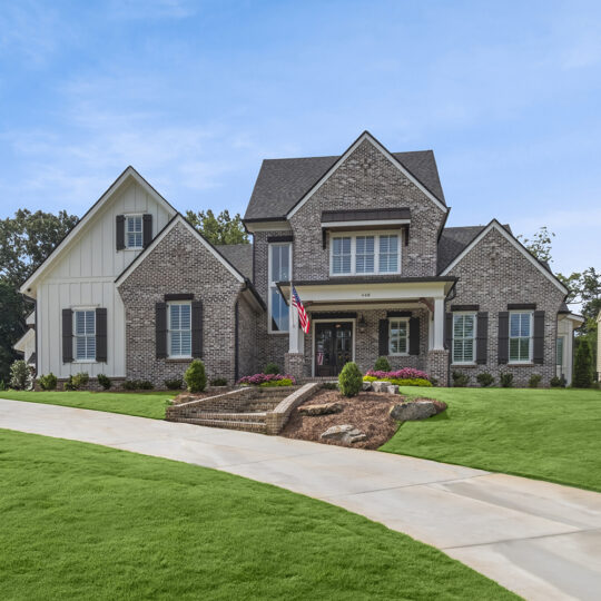 A two-story house with a mix of brick and light siding, featuring a triangular roof and a neatly landscaped front yard. A long, curved driveway leads to the entrance. An American flag is displayed near the steps.