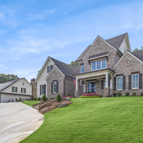 Large brick house with gabled roof, American flag by the entrance, and manicured lawn. A curved driveway leads to a detached garage. The sky is clear and blue, with trees in the background.